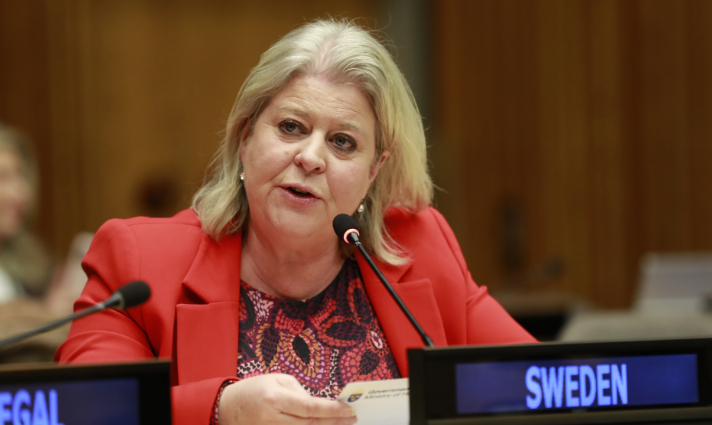 Minister for Social Affairs Camilla Waltersson Grönvall spekling into a microphone. In front on her is a sign spelling out "Sweden". 