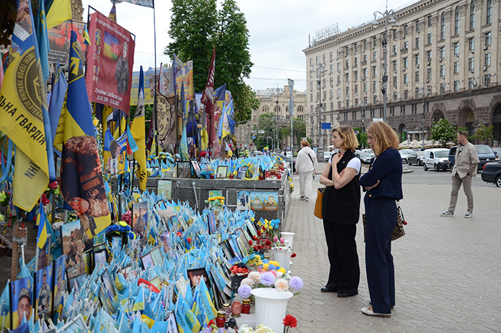 Minister for Gender Equality Nina Larsson and Minister for Health Care Elisabet Lann stand at a memorial site in Ukraine, viewing rows of small Ukrainian flags, photographs, flowers and banners commemorating fallen soldiers, with buildings and traffic in the background.