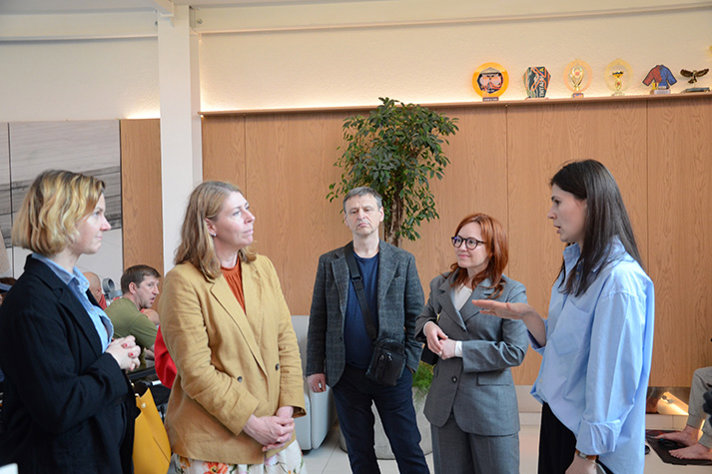 Minister for Health Care Elisabet Lann and Minister for Gender Equality Nina Larsson stand with several people inside a rehabilitation facility at the Superhumans Center, listening during a discussion, with a seating area, a potted plant and decorative items visible in the background.