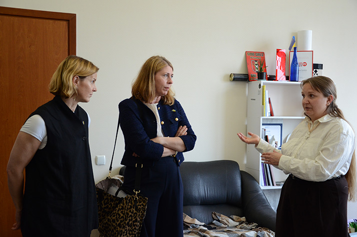Minister for Health Care Elisabet Lann and Minister for Gender Equality Nina Larsson stand and speak with a representative inside an office at the Ukrainian Women Veterans Movement (Veteranka), with a sofa, a shelf with books and objects, and a door visible in the background.