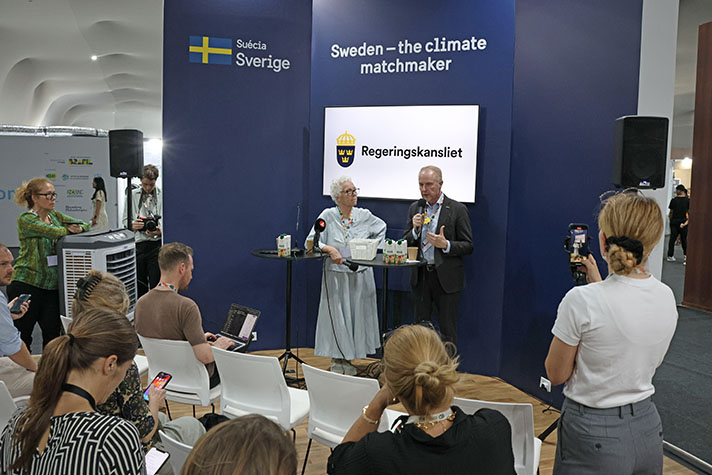 The two persons are standing at a high table in front of a blue wall.