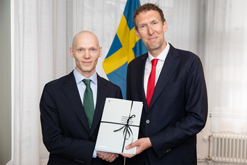 Two men in suits looking facing the camera, holding papers and shaking hands. Swedish flag in background. Indoors.