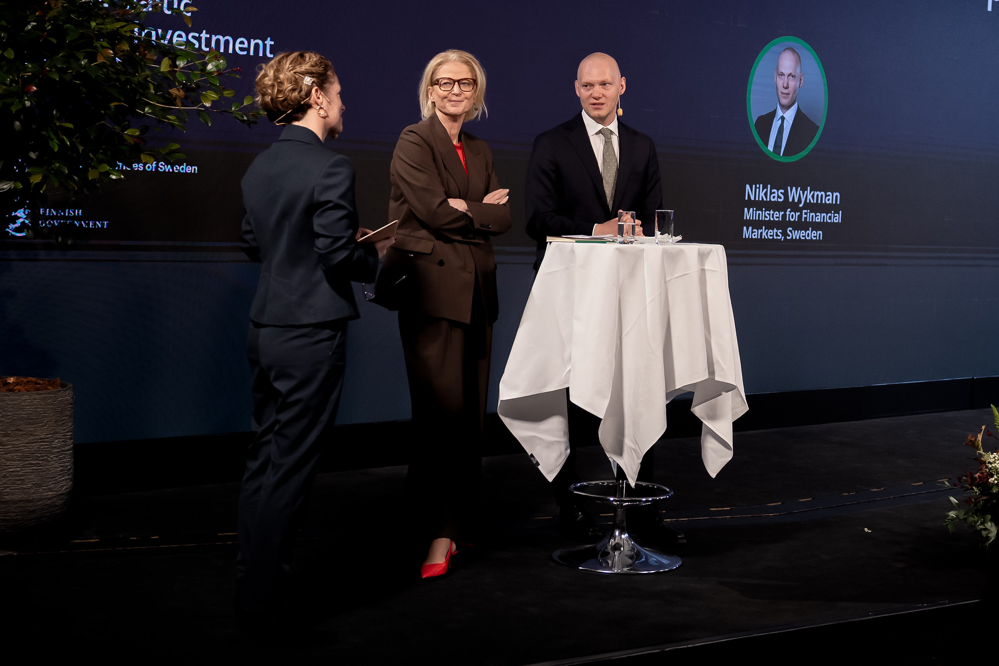Two ministers in a discussion with the moderator. They are standing close together on the scen.