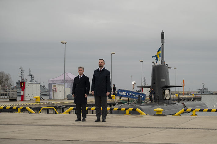 Pål Jonson and Władysław Kosiniak-Kamysz stands at attention by a dock in Gdynia. It's a cloudy day and in the background the swedish submarine HMS Uppland is docked.