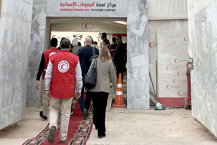 People enter a humanitarian aid‑packing centre, guided by staff in red vests bearing the Red Crescent emblem. The entrance is decorated with banners and photographs, and a red carpet covers the ground.