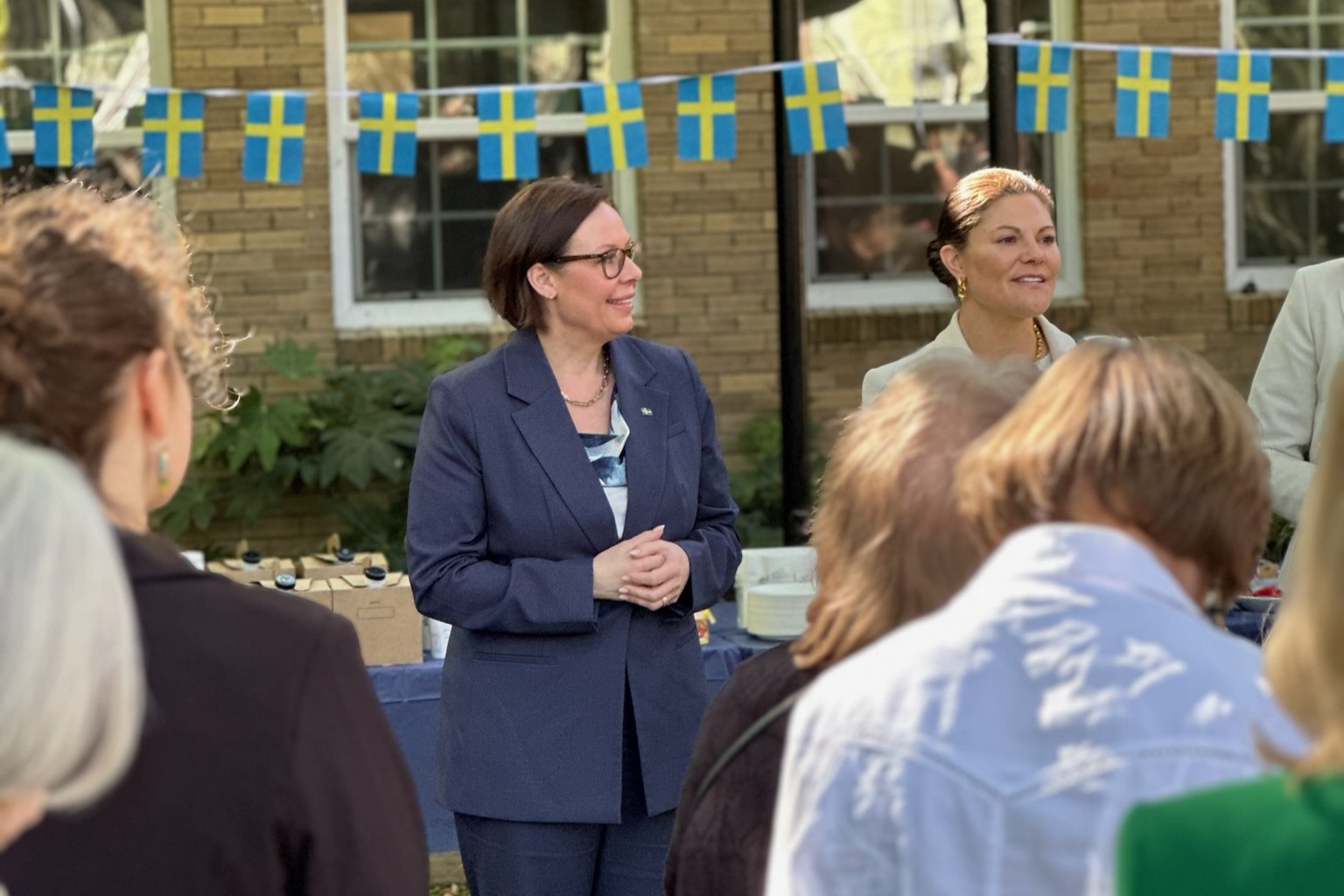 The Minister for Foreign Affairs and the Crown Princess standing outside in front of a garland with Swedish flags.
